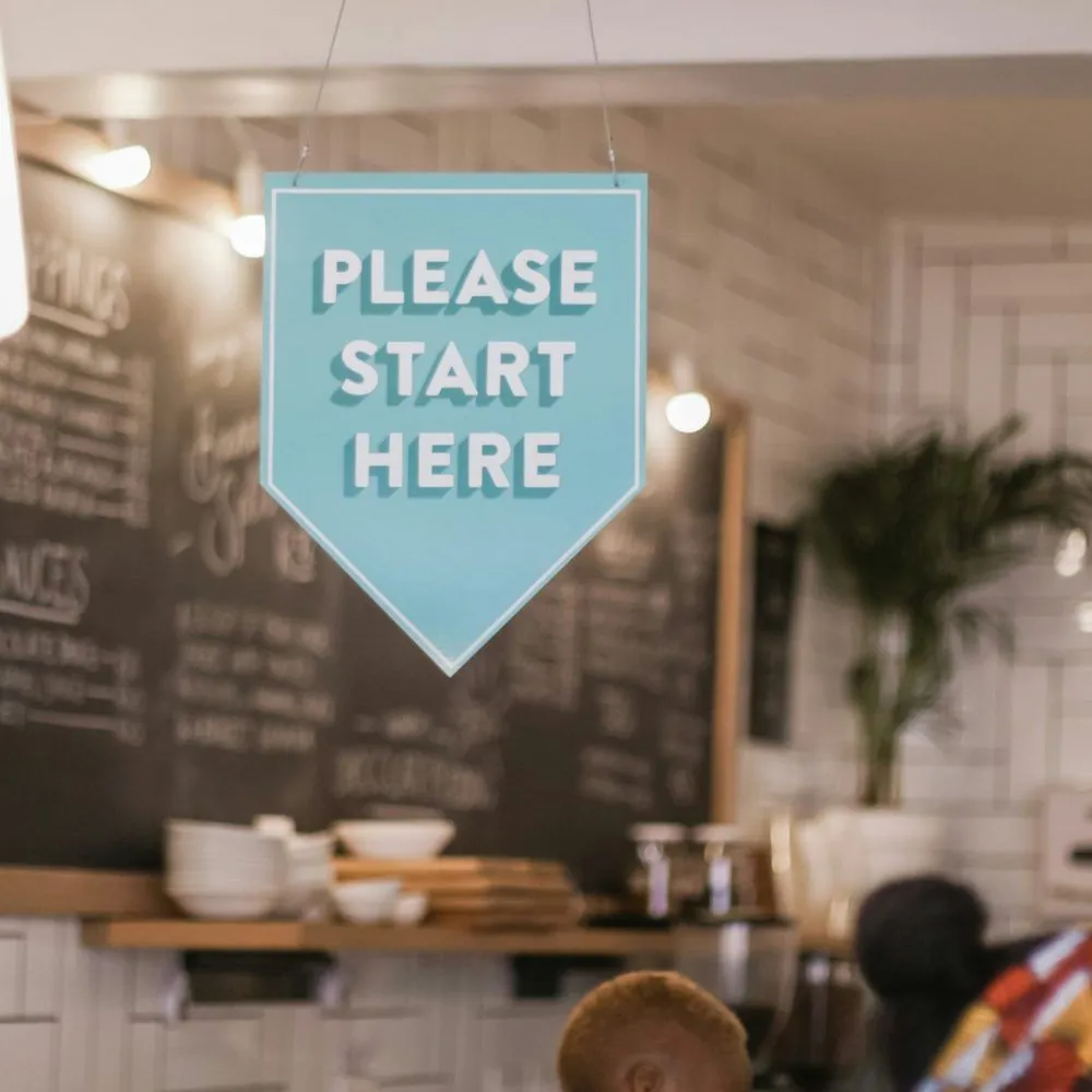 Café-Hintergrund unscharf, Schild „Please start here“ sichtbar.