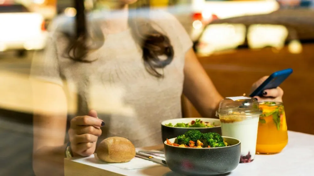 Frau sitzt am Tisch mit Essen und schaut auf ihr Smartphone.