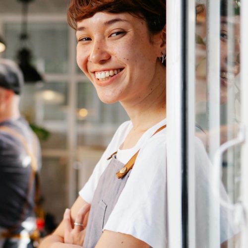 Kellnerin, die an einer Fensterscheibe lehnt und in die Kamera lächelt