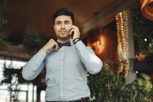 Mann mit Hemd und Fliege hält sich ein Smartphone an sein Ohr